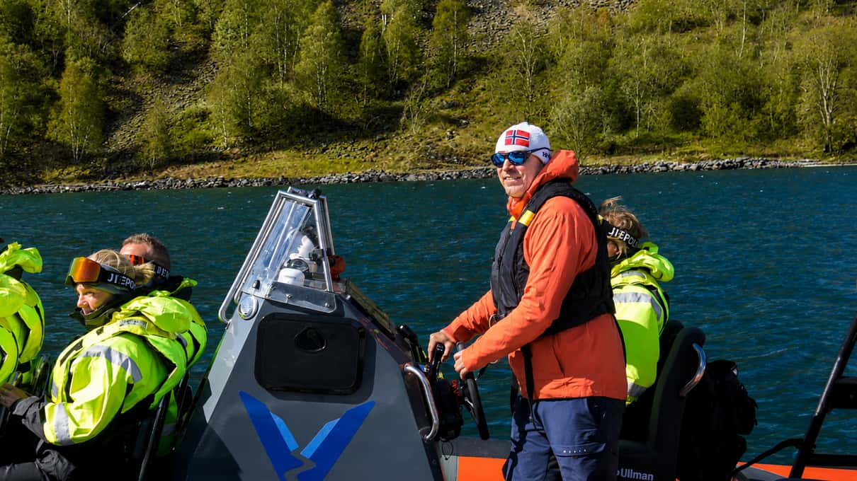 A guide standing at the console of a RIB boat while passengers in life jackets watch the steep green fjord wall behind
