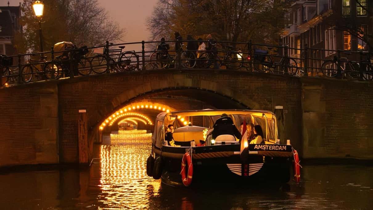 Offenes Boot mit Gästen fährt in der Abenddämmerung auf eine mit Lichterketten beleuchtete Brücke zu