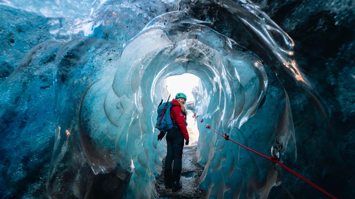 Visitor with a red backpack standing inside a curving blue ice cave tunnel under Vatnajökull glacier