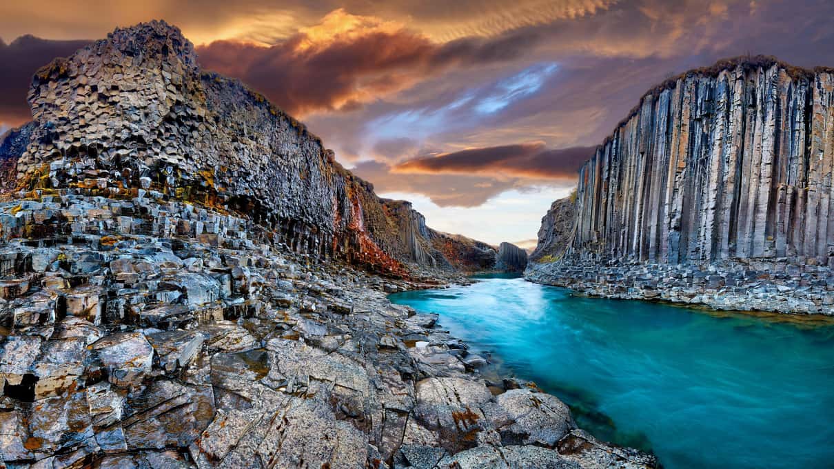 Basalt columns lining a turquoise river in the canyon under an orange and red sunset sky