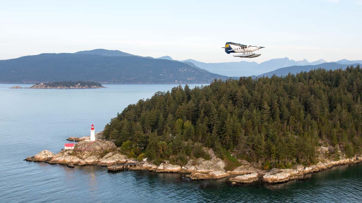 Floatplane flying past the forested point of Lighthouse Park with its red and white lighthouse on the rocks