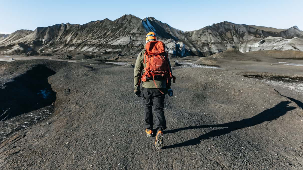 En turgåer med rød sekk på mørk grus under fjellryggene nær Mýrdalsjökull på en tur med Troll.is