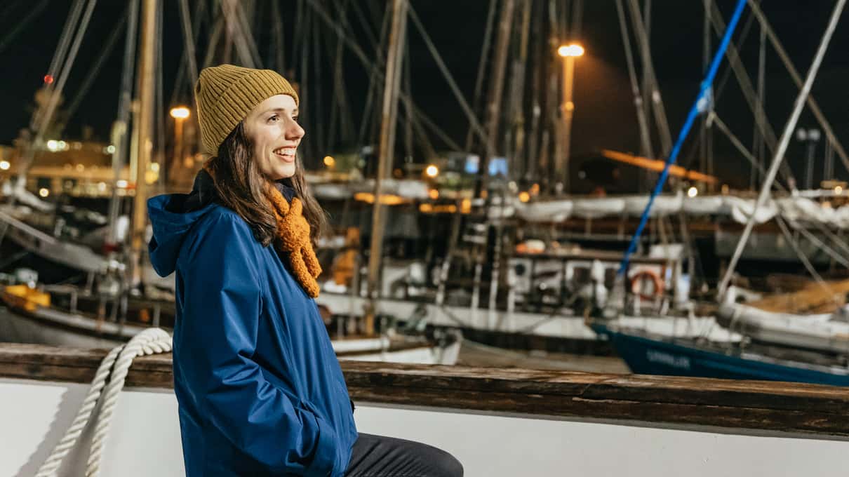 A woman in a beanie standing at the Reykjavik marina at night with boat masts behind her