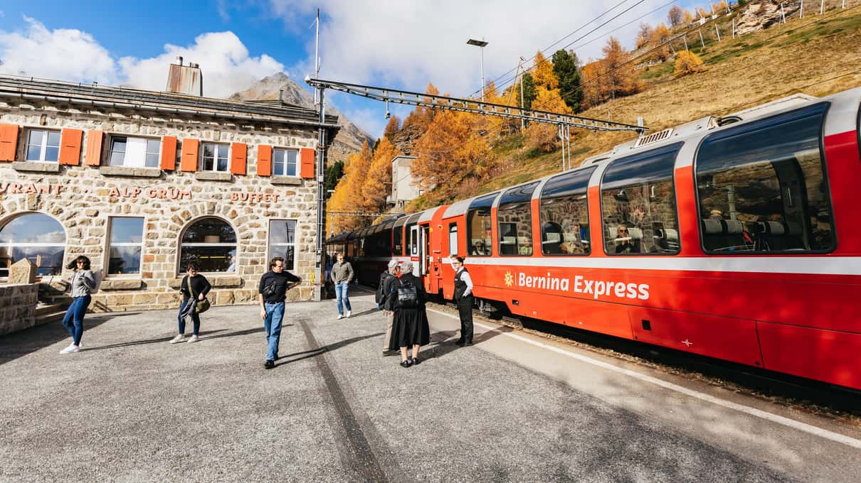 Travelers standing outside a mountain station building beside the red Bernina train