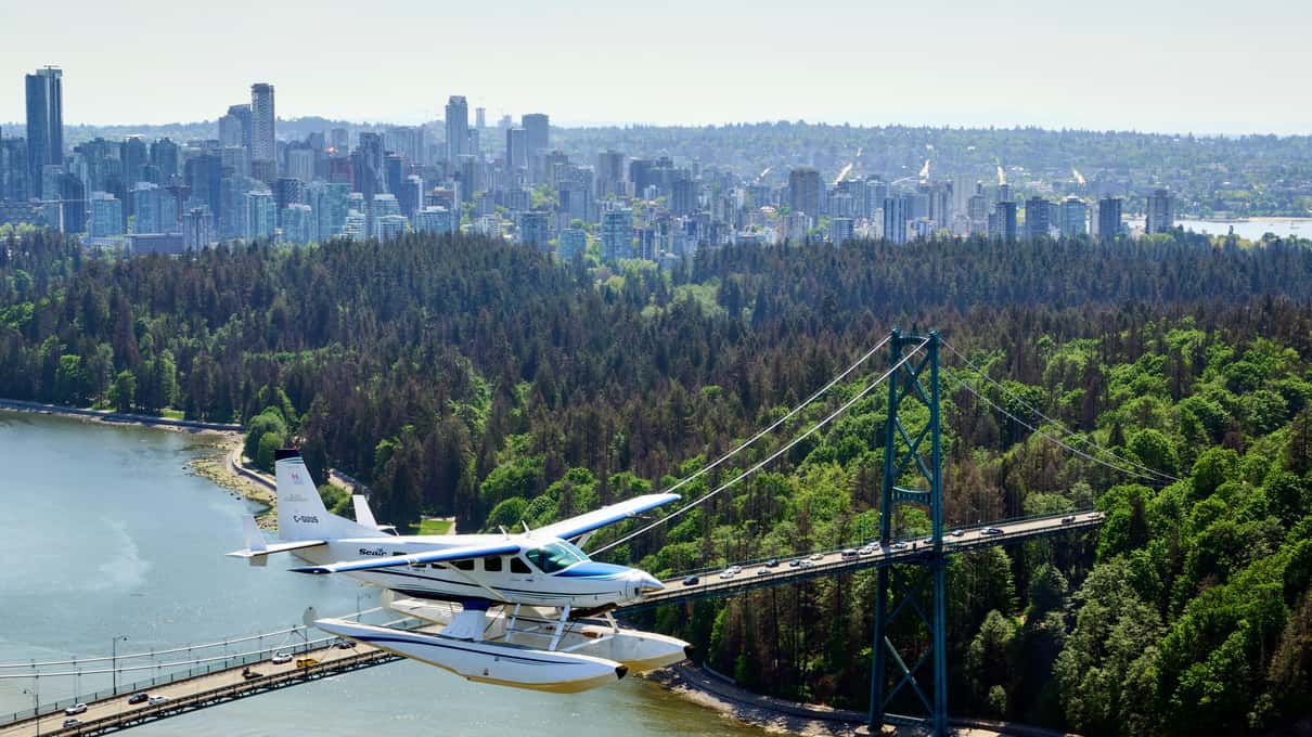 A floatplane flying low past the green towers of the Lions Gate Bridge with Stanley Park and downtown Vancouver behind