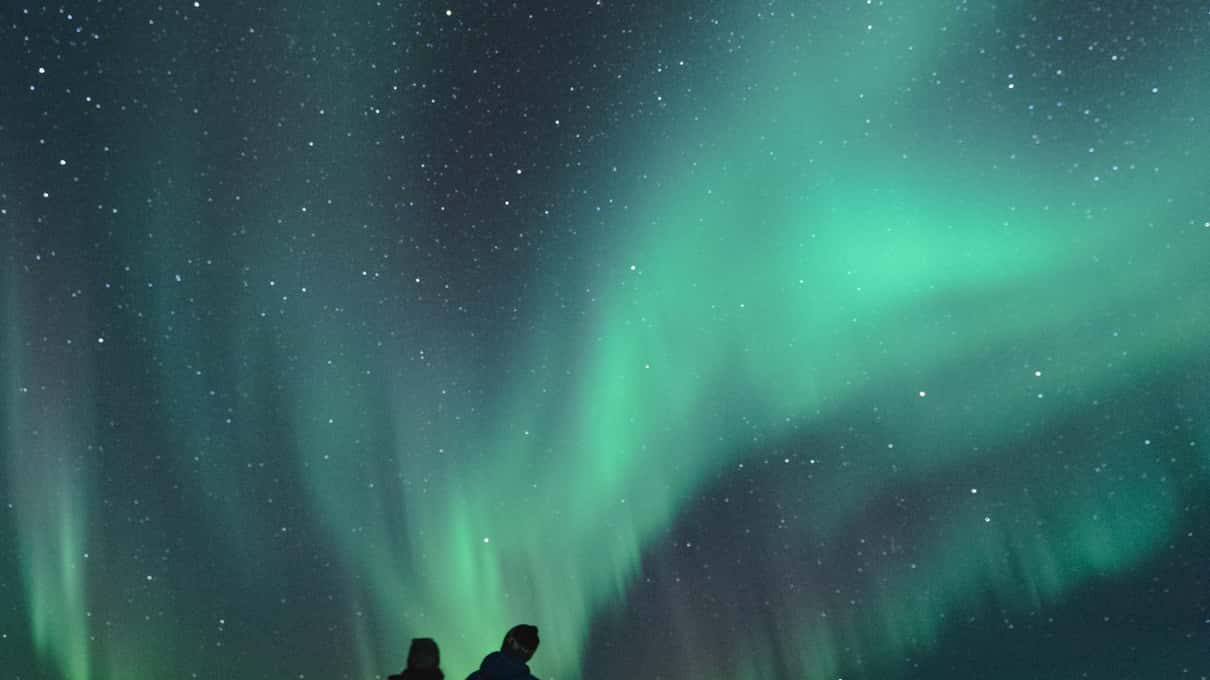 Two people standing small at the bottom of the frame under a wide green aurora filling the starry sky over Lofoten