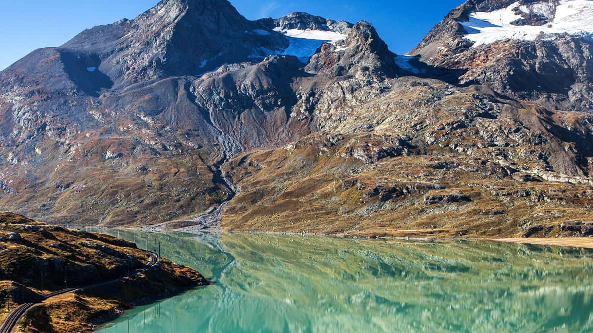 The red Bernina train running along the shore of the turquoise Lago Bianco below bare mountain slopes