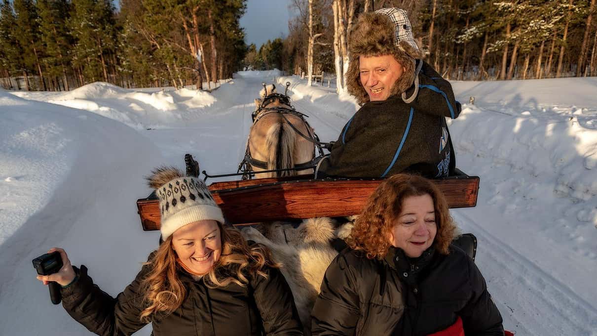 Two smiling guests in fur hats wrapped in thick blankets on a horse sleigh in the snow near Alta