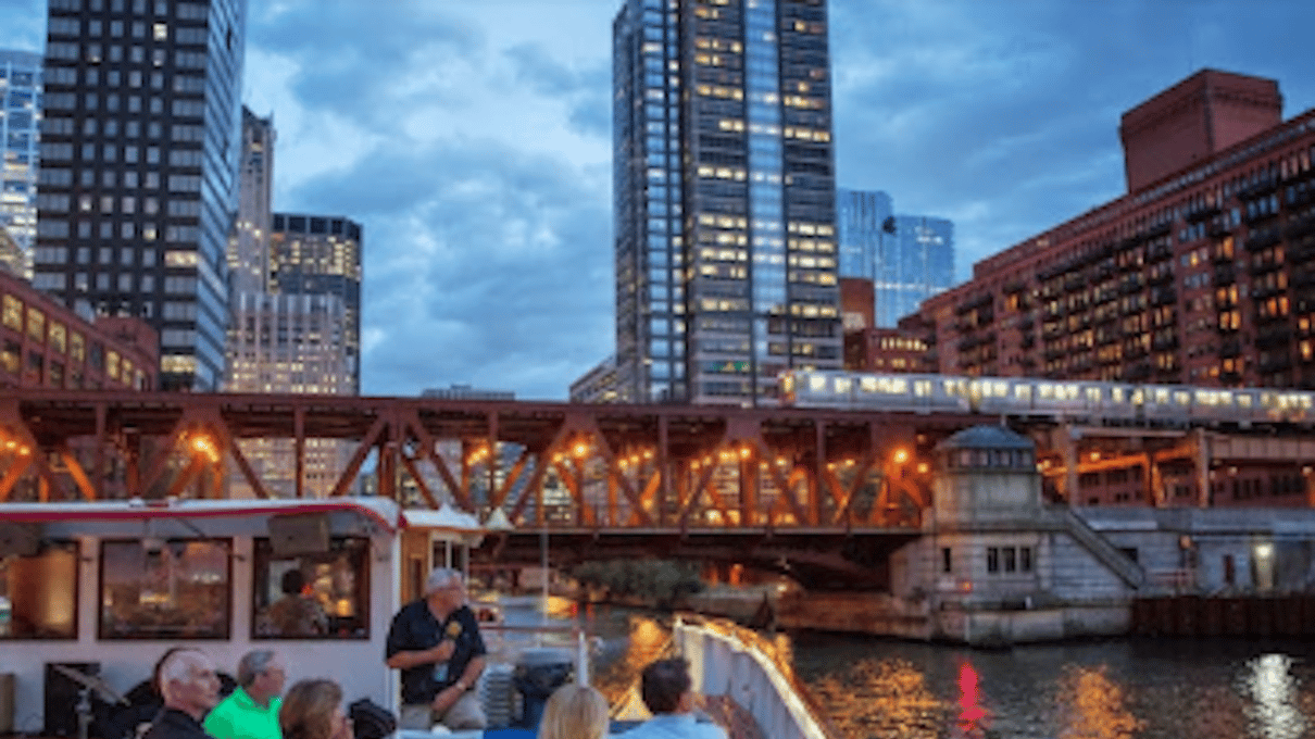 River cruise boat passing under a lit Chicago bridge in the evening with towers behind