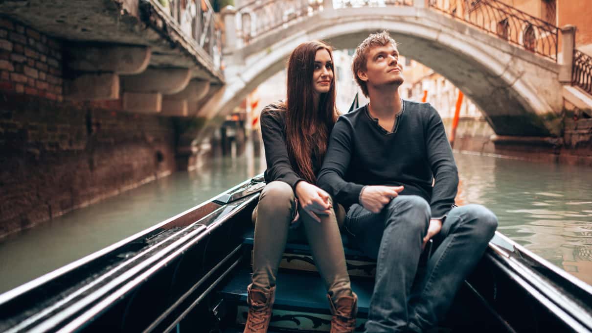 Pareja sentada en la proa de una góndola en un canal de Venecia, mirando a lo lejos, con un puente de piedra detrás