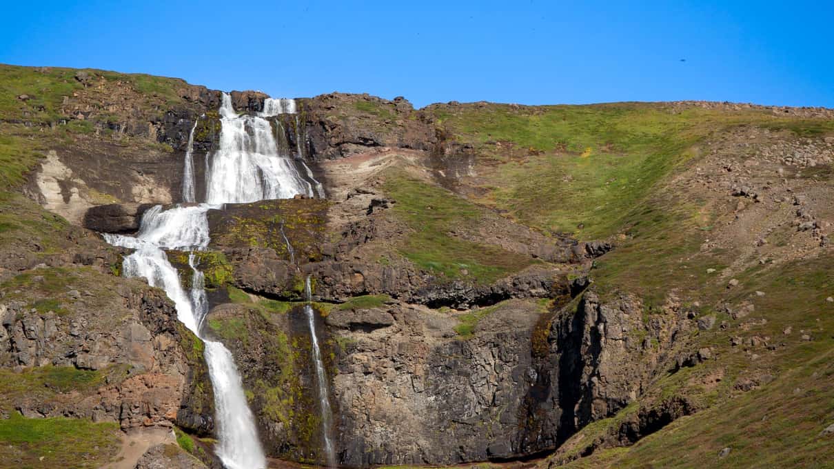 A tall waterfall falling in several tiers down a steep green hillside in East Iceland