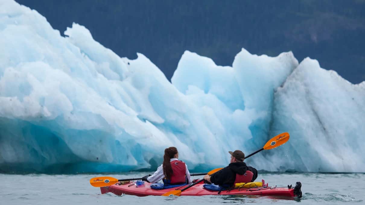 Two paddlers in a red tandem sea kayak passing a wall of blue and white iceberg ice