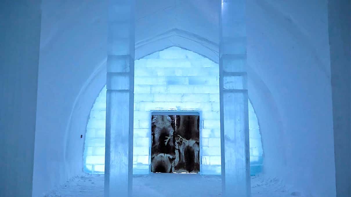 A corridor of ice pillars leading to a lit doorway with reindeer-hide doors inside the Icehotel