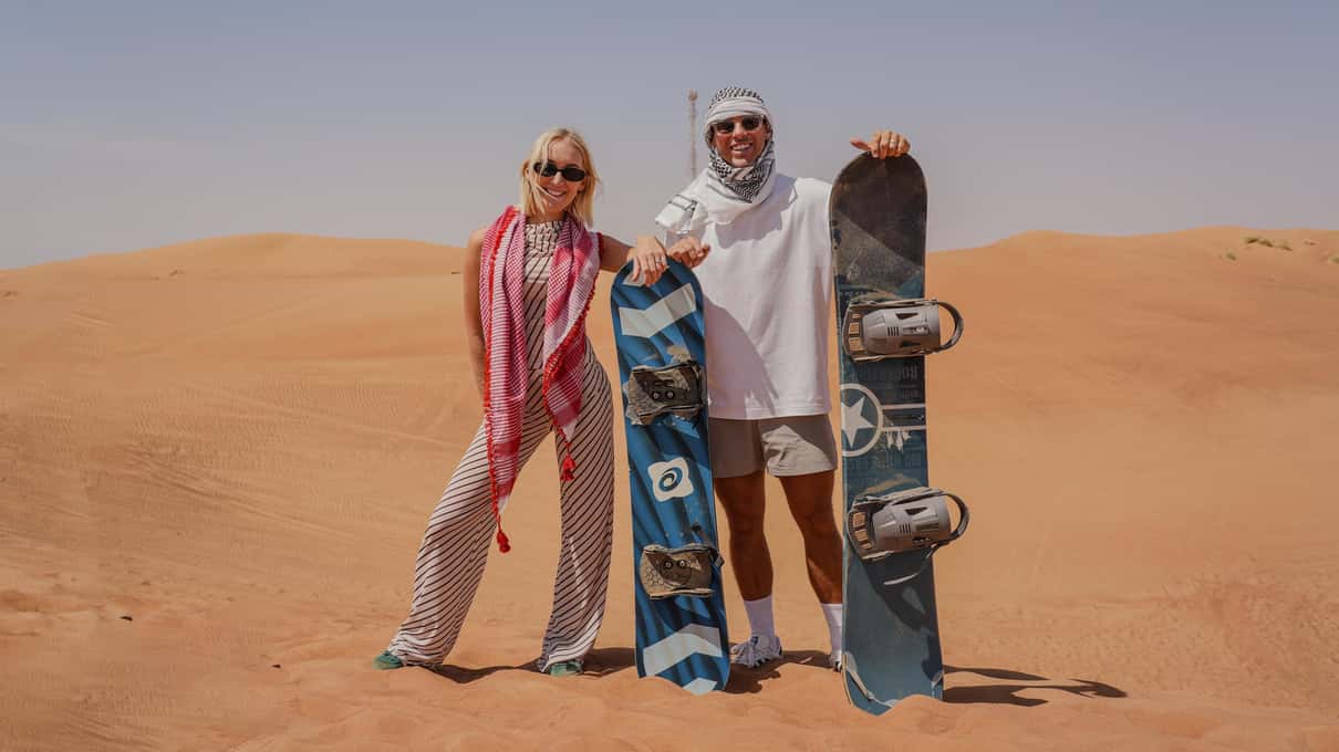 Pareja con pañuelos posa sonriente en las dunas de Dubái con dos tablas de sandboard
