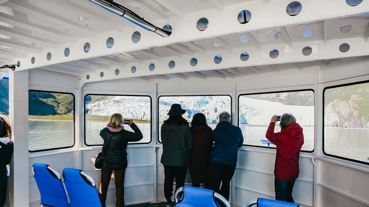 Passagerer i varme jakker med kikkerter ved vinduerne i en opvarmet bådkabine med udsigt mod en gletsjer