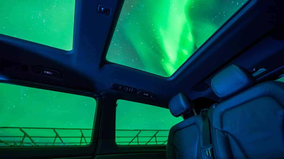 A green aurora seen through the panoramic glass roof of a Mercedes van, seats in the foreground