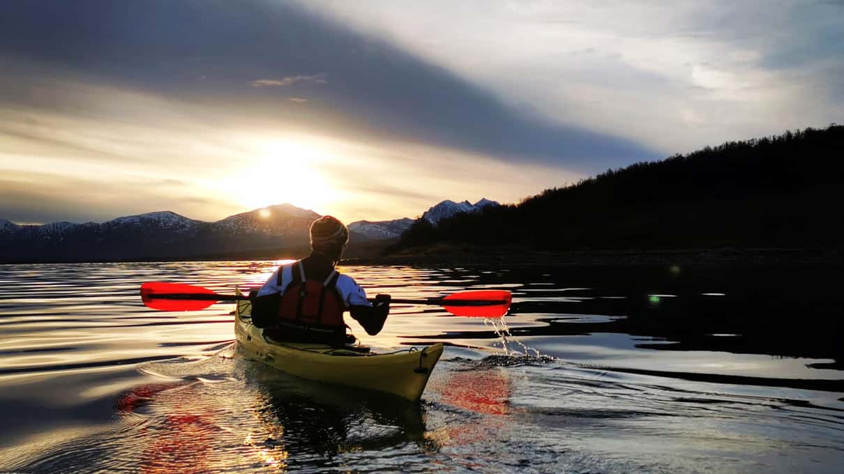 A kayaker paddling a yellow sea kayak toward a low winter sun between dark hills