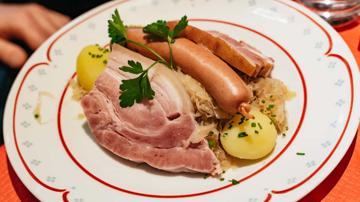 A plate of choucroute garnie with sauerkraut, smoked pork, two sausages and boiled potatoes on a red-rimmed plate