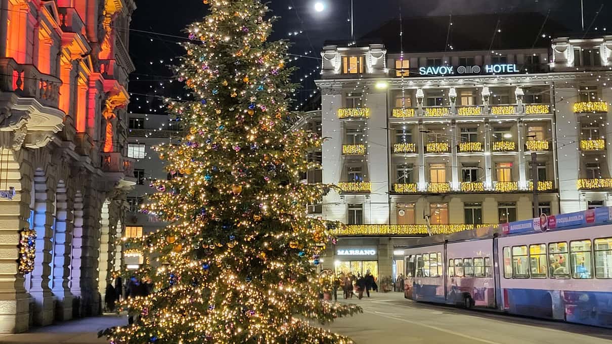 A tall Christmas tree covered in warm lights on Paradeplatz at night, with a blue Zurich tram and the lit Savoy hotel behind