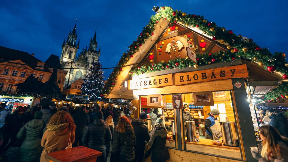 A lit wooden klobása sausage hut at a Prague Christmas market at blue hour, with Gothic spires rising against the evening sky behind it