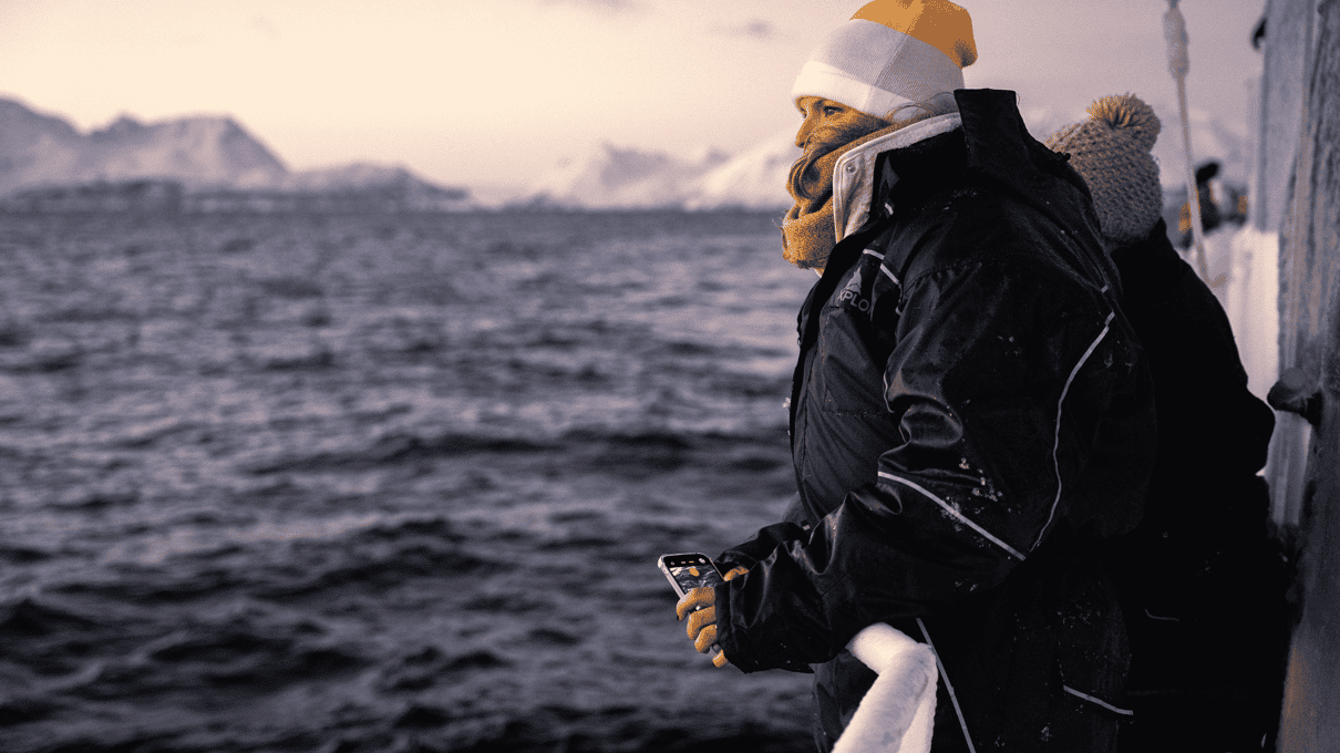 A guest in a warm beanie at the rail of Polar Adventures' boat, looking over the sea under a pink Arctic sky