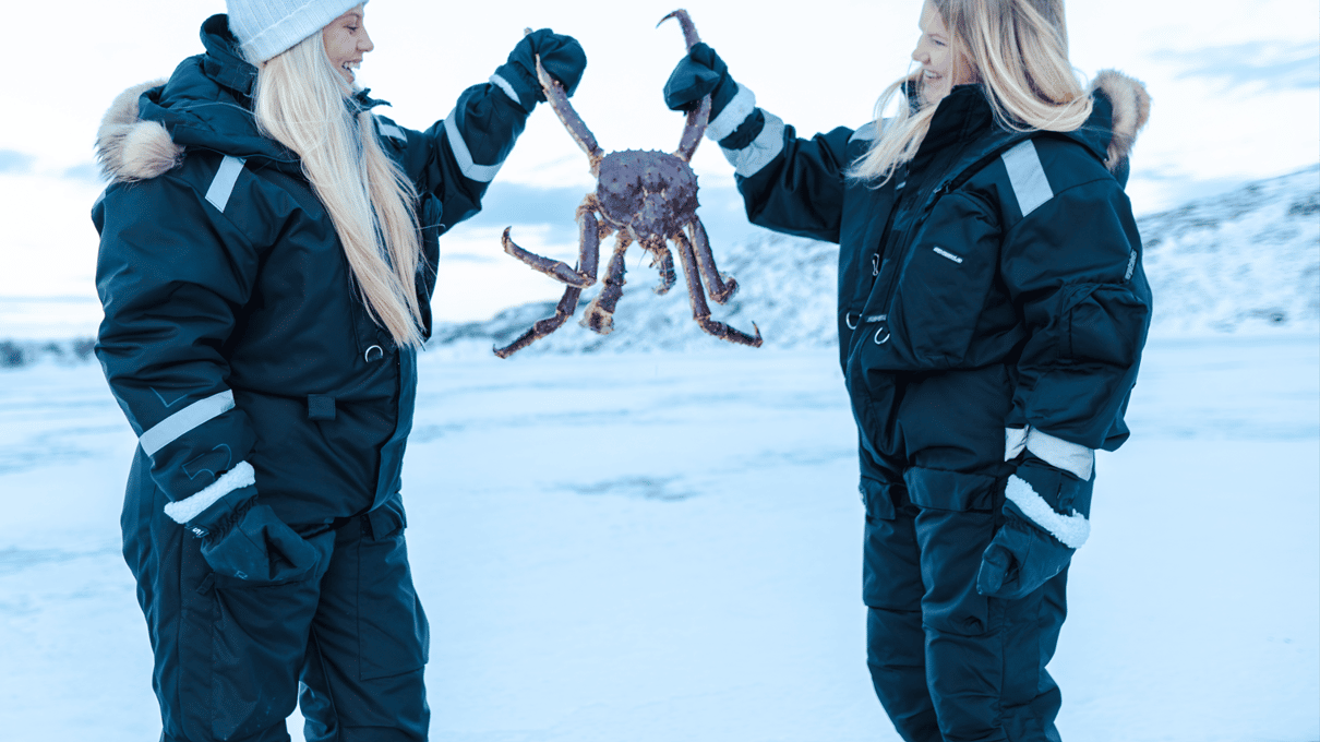 Two guests in thermal suits holding a red king crab between them on the frozen fjord near Kirkenes