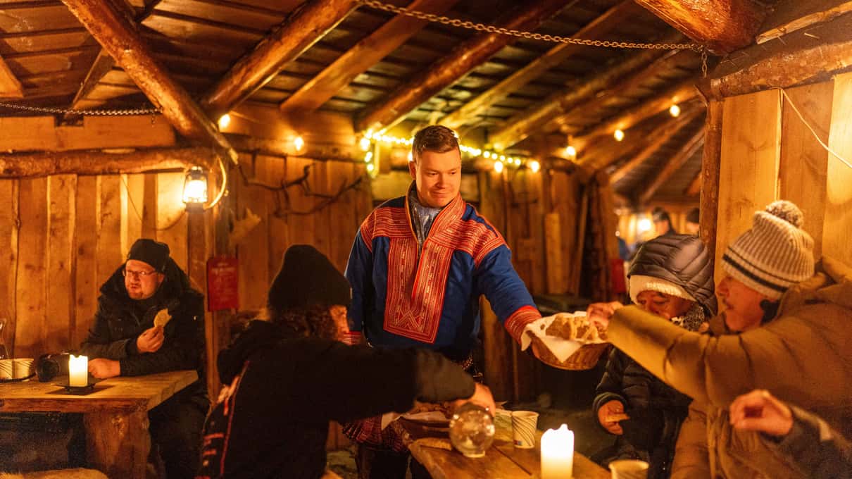 A Sami host in a traditional blue tunic serving guests at candle-lit wooden tables inside a turf hut