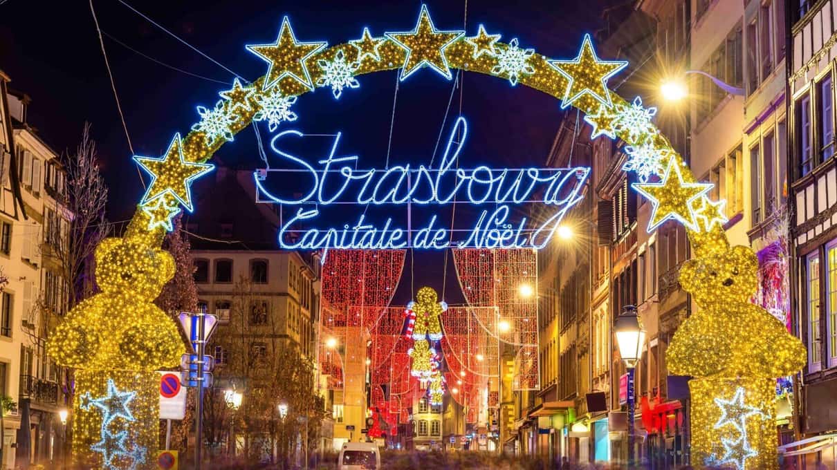 A star-shaped light arch reading Strasbourg Capitale de Noël with two gold teddy bears above a lit street at night
