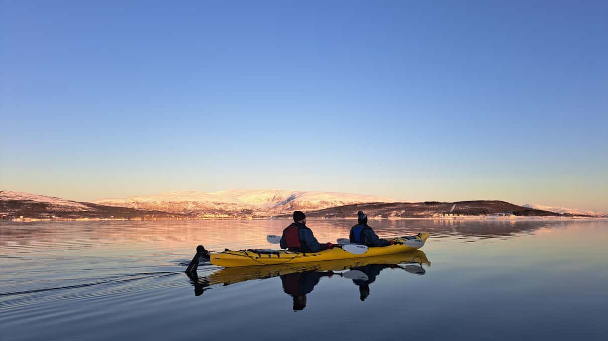 A yellow double kayak crossing calm water below a coast lit pink by the low winter sun