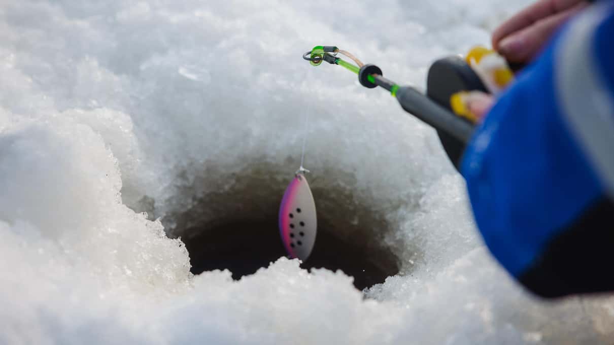 A short ice rod lowering a spotted lure into a deep, narrow hole drilled through snow and ice