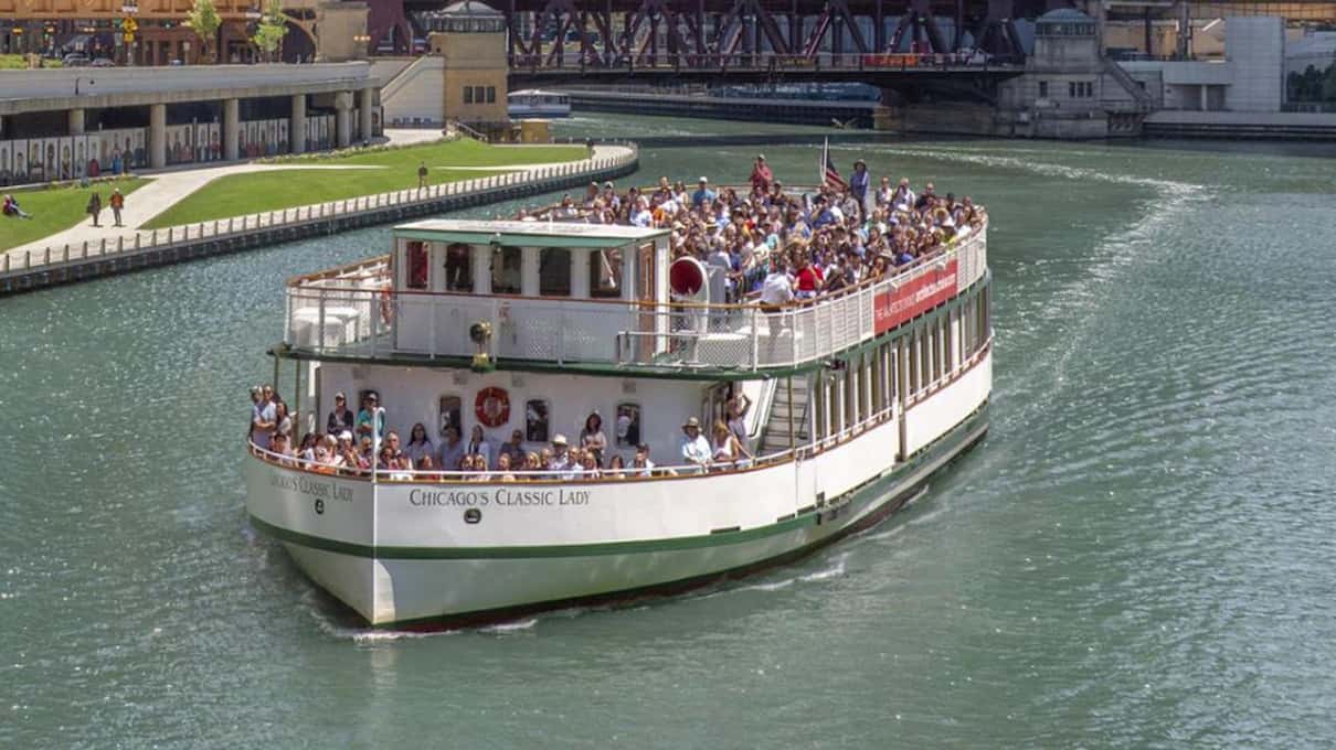 A white river cruise boat with a full upper deck of passengers sailing along the Chicago River