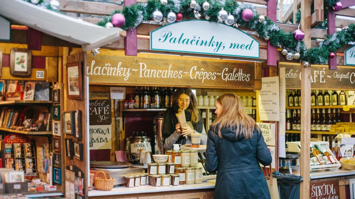 A Prague Christmas market stall selling gingerbread and honey, with a woman browsing jars and decorated biscuits