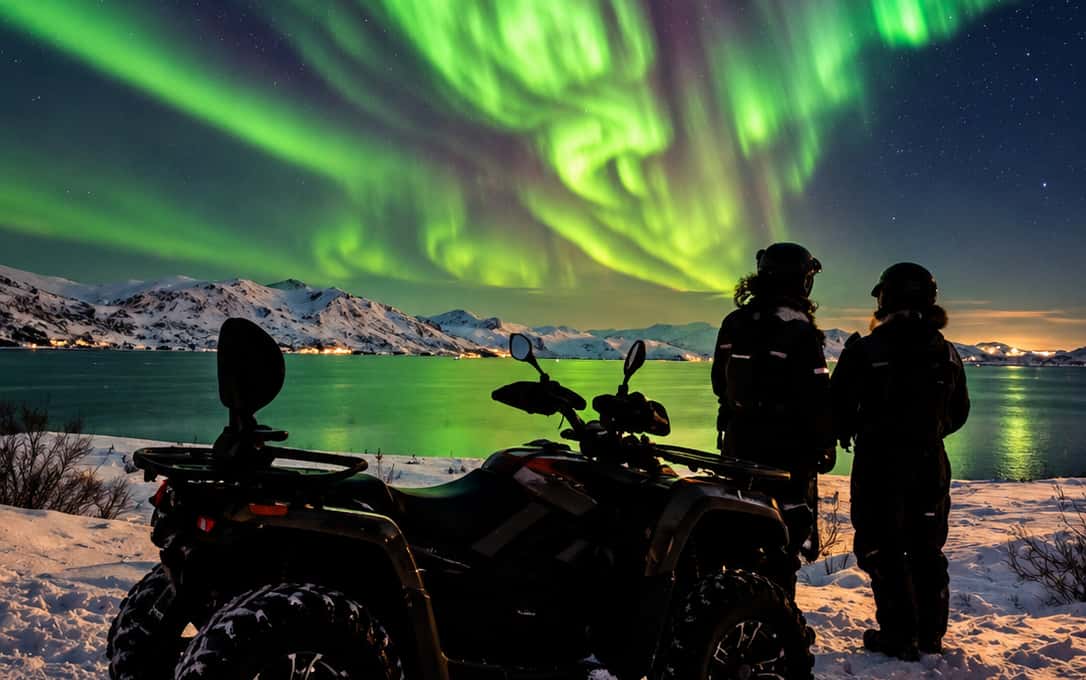 Two riders beside a parked quad watching a green aurora over a fjord with lights along the shore