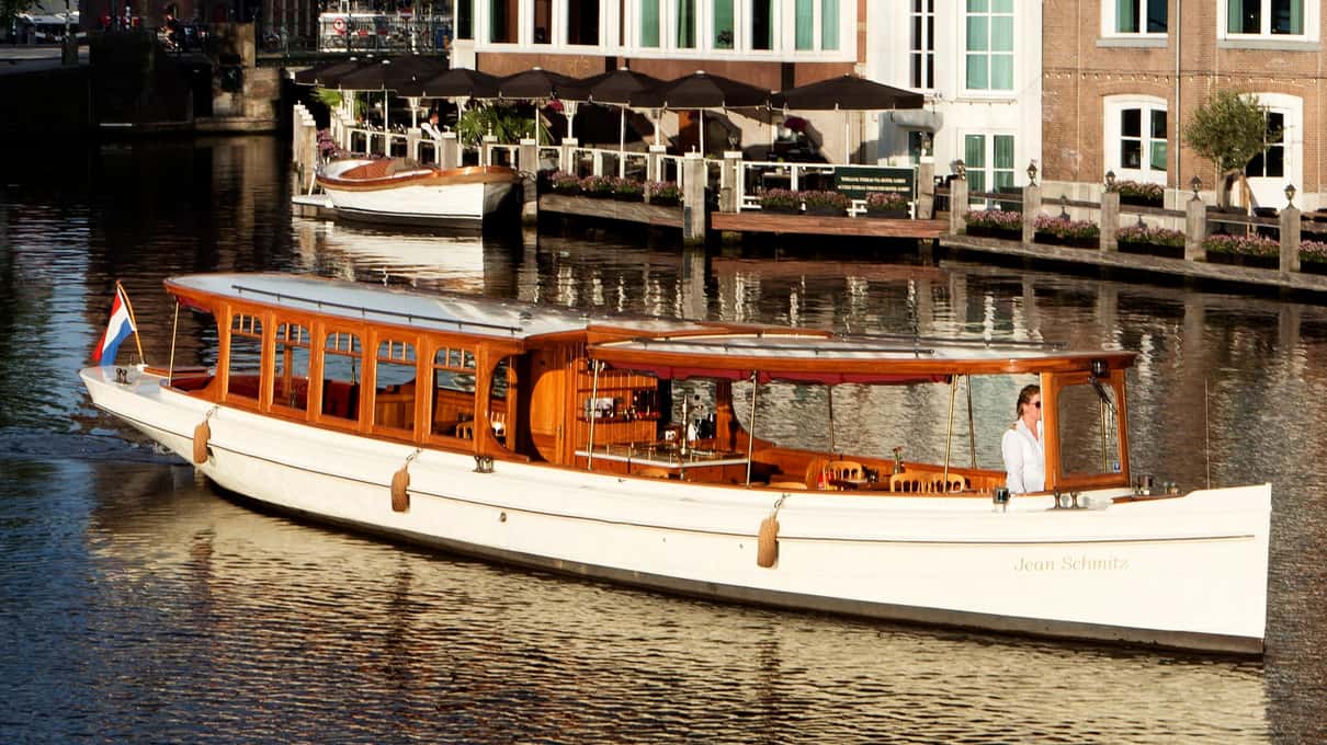 Klassisches Salonboot mit weißem Rumpf und Holzaufbau liegt an einer Gracht vor Häusern