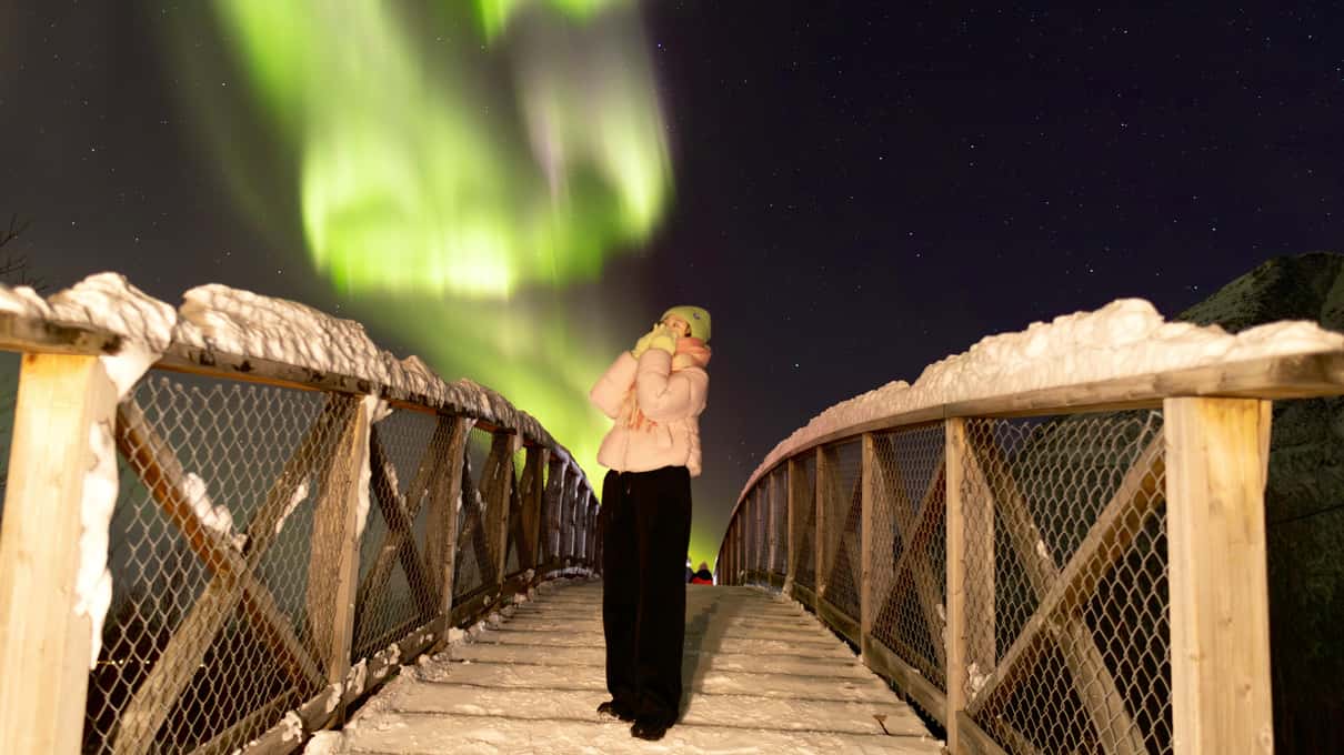 Una mujer de pie en una pasarela de madera cubierta de nieve, de noche, bajo una aurora verde
