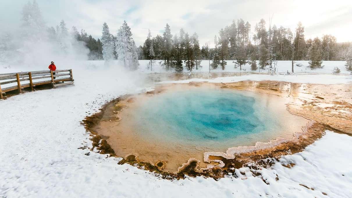 A steaming hot spring with a deep blue center and an orange rim set in thick snow