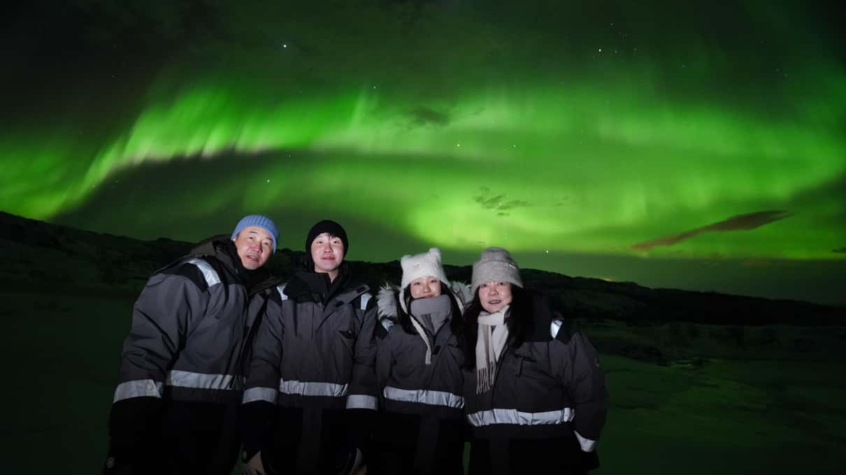 Vier Gäste in grauen Thermoanzügen stehen nachts nebeneinander, hinter ihnen ein breites grünes Nordlicht