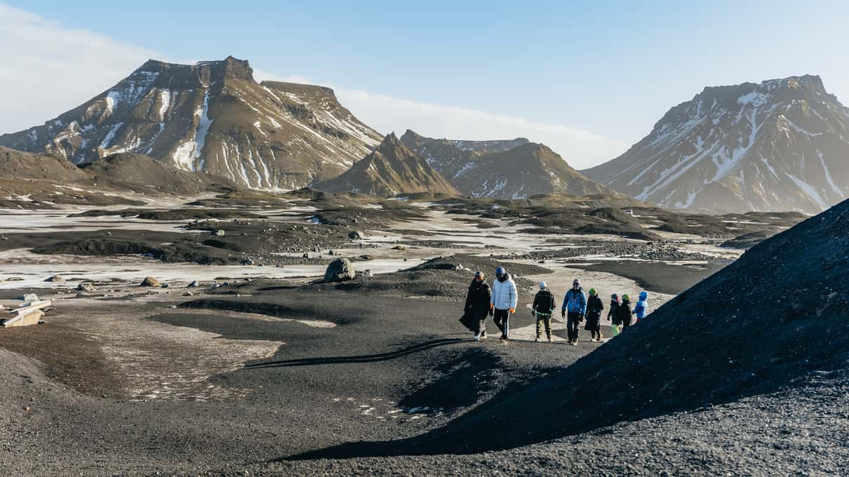 A small group walking across black volcanic sand toward the mountains near the glacier