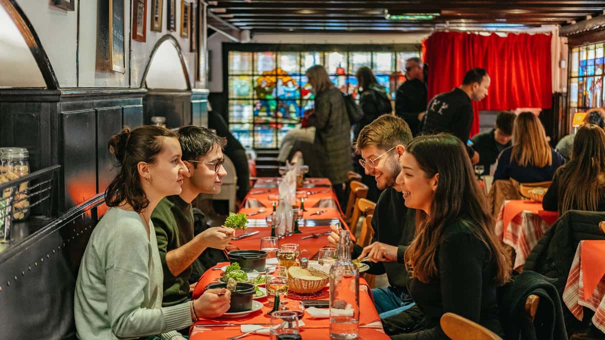 A food tour group seated along a long table with a red cloth in a wood-panelled Alsatian tavern, plates and wine glasses in front of them