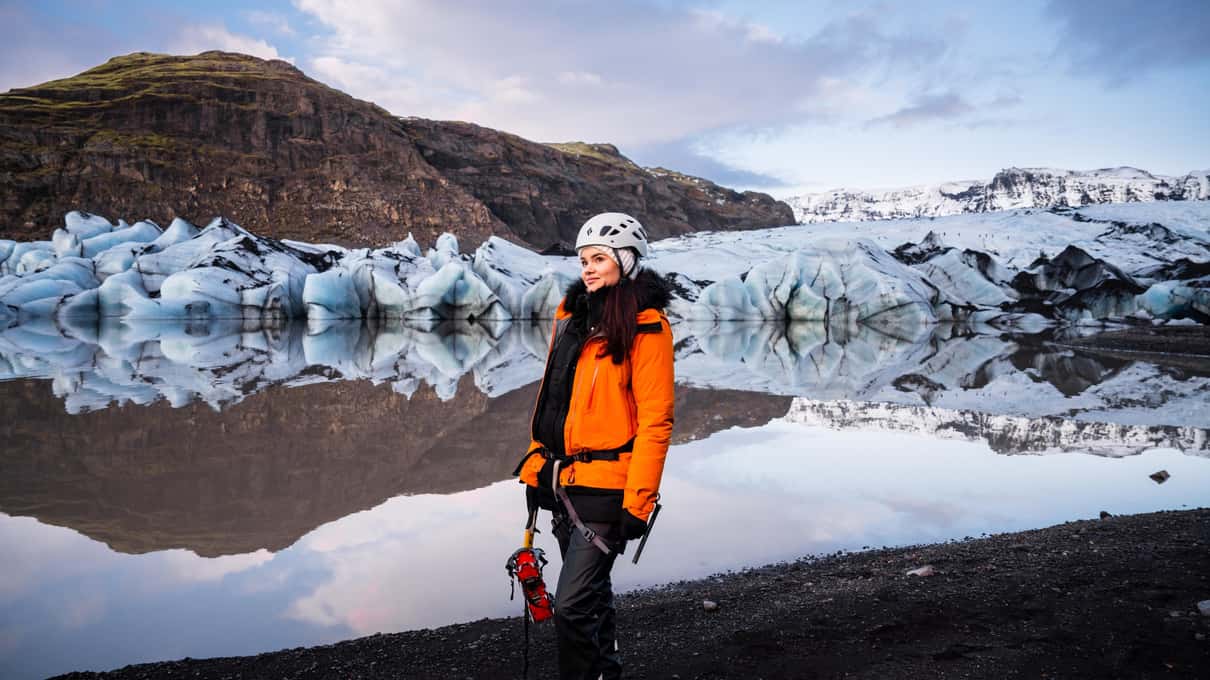 A hiker in an orange jacket standing by the Sólheimajökull glacier lagoon with floating ice behind