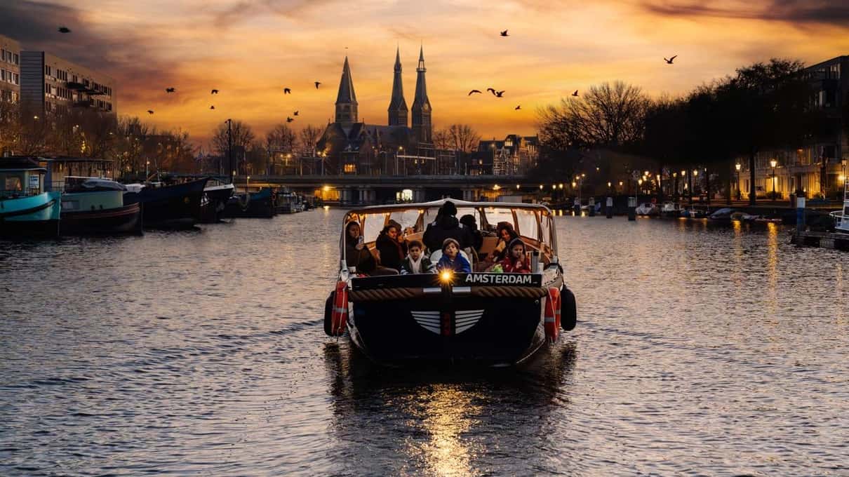 Open rondvaartboot op een Amsterdamse gracht bij zonsondergang, met de torens van de Westerkerk tegen een oranje lucht