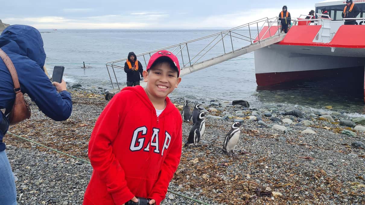 A visitor in a red jacket on the Magdalena Island shore with a red-and-white ship moored behind