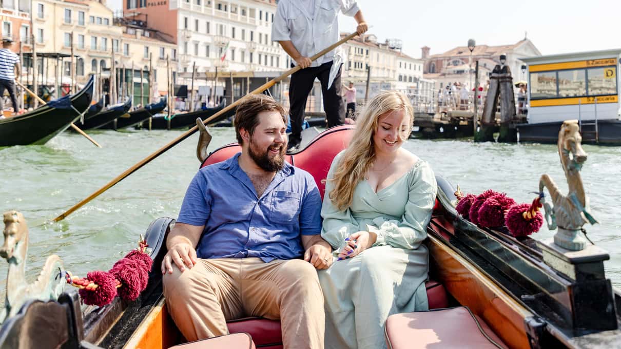 Stel zit lachend op de rode kussens van een gondel op het Canal Grande, achter hen roeit de gondelier
