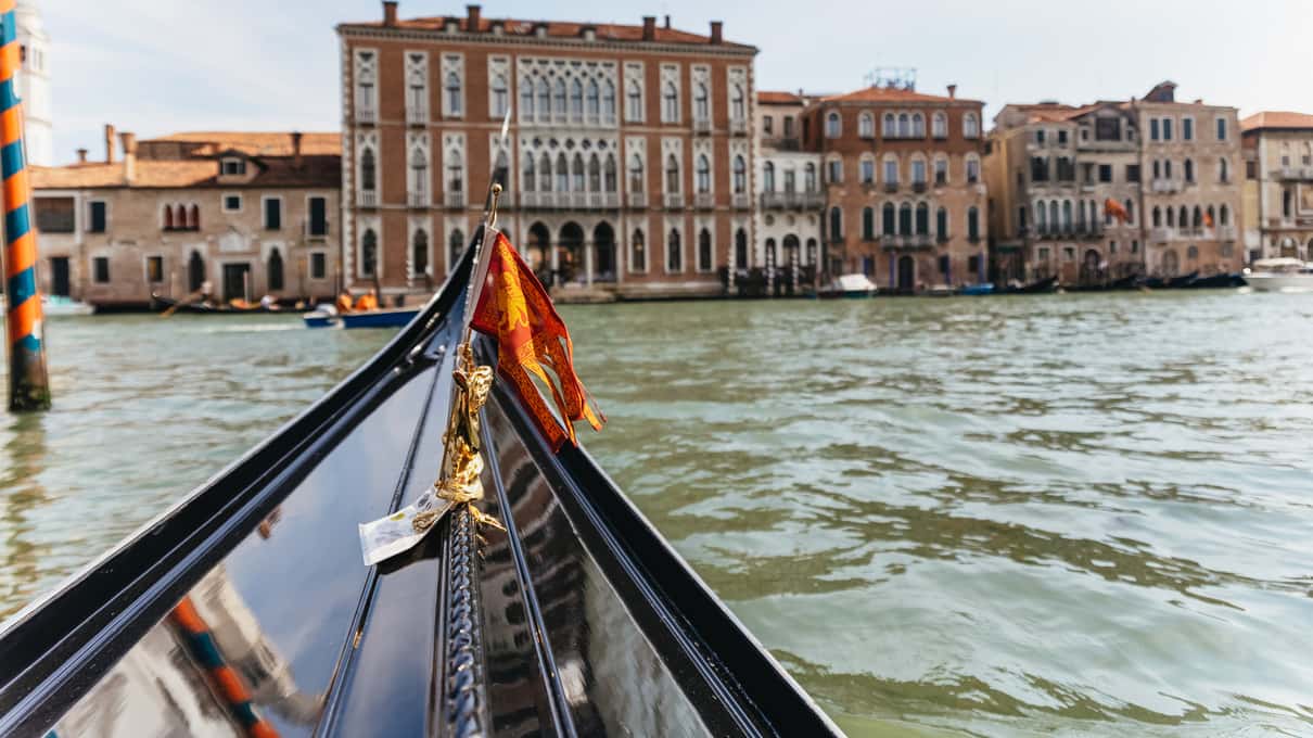 Proa negra de una góndola con el hierro plateado y la bandera de Venecia avanzando por el Gran Canal entre palacios
