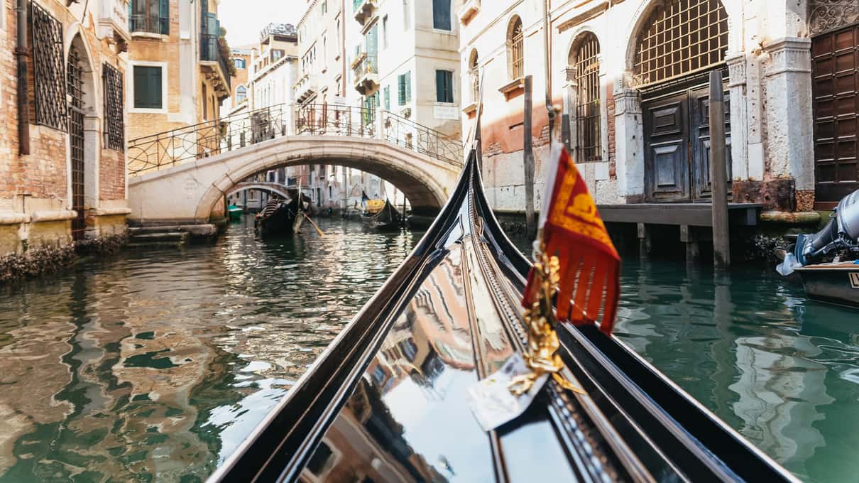 Blick über den schwarzen Bug einer Gondel mit venezianischer Flagge in einen schmalen Kanal mit Steinbrücke