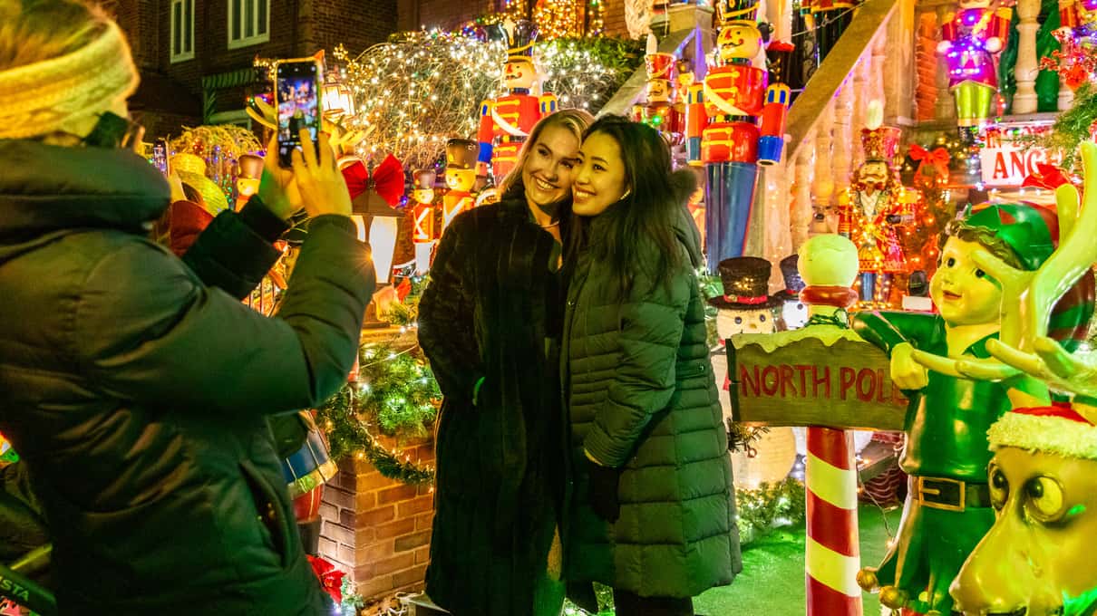 Two friends in winter coats and knitted hats taking a selfie beside lit Christmas figures in Dyker Heights
