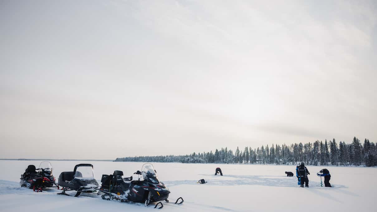 Snowmobiles parked on a vast frozen lake near the Arctic Circle with ice anglers in the distance