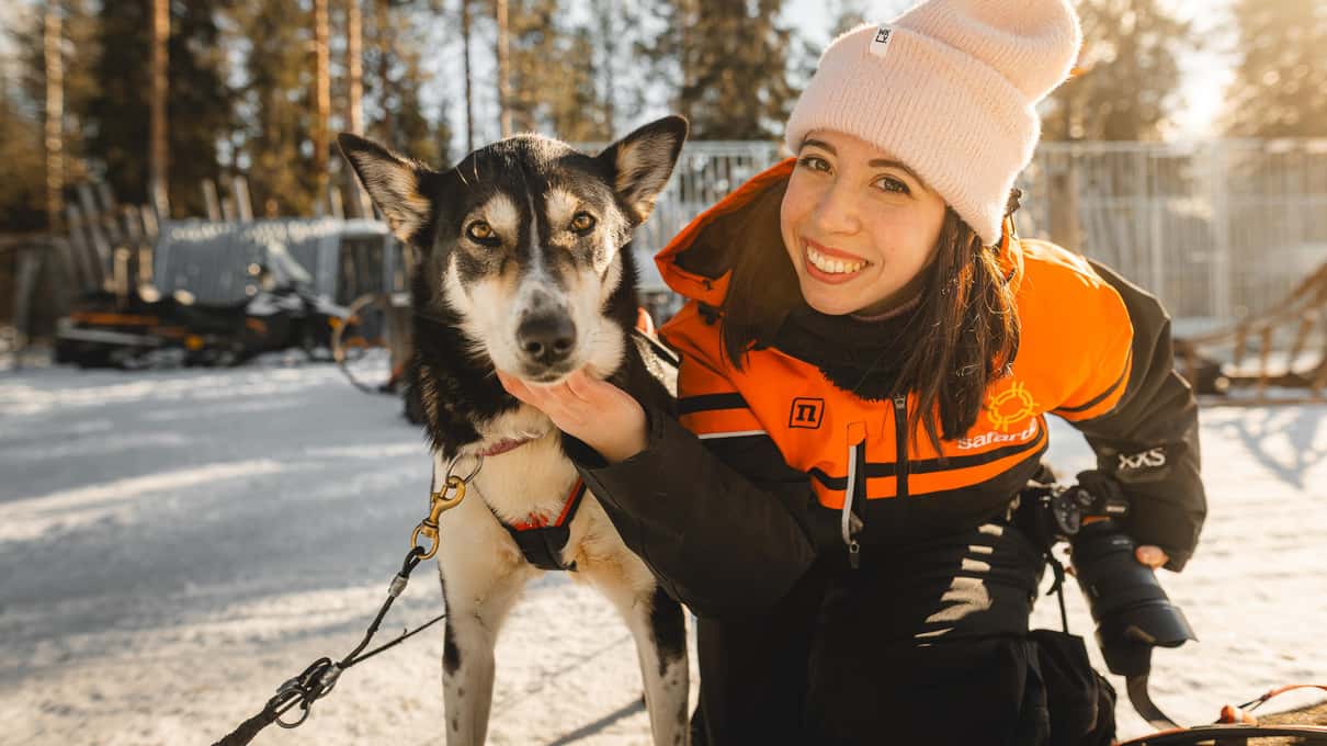 A smiling guest in an orange winter suit and hat hugging a husky at a Lapland kennel