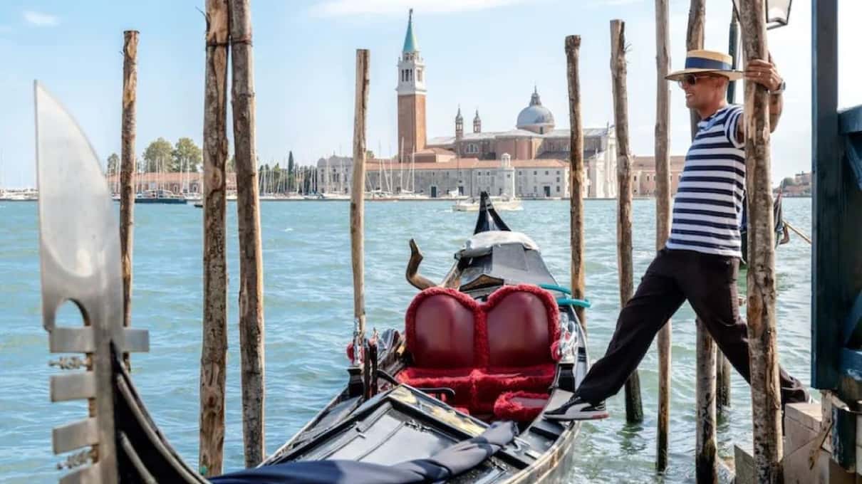 Gondoliere in maglia a righe sale su una gondola dai cuscini rossi tra le paline, sullo sfondo il campanile di San Giorgio Maggiore
