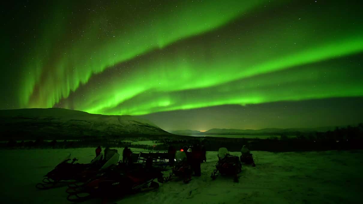 Bright green aurora bands above a group of riders and parked snowmobiles on the snow